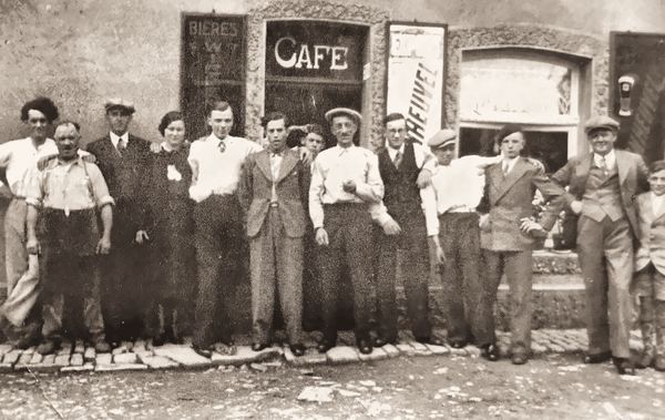 Rassemblement plus large, les hommes du quartier. La mode d'avant-guerre est évidente : bérets, casquettes, bretelles, vestons… Ils ont fière allure. Un…